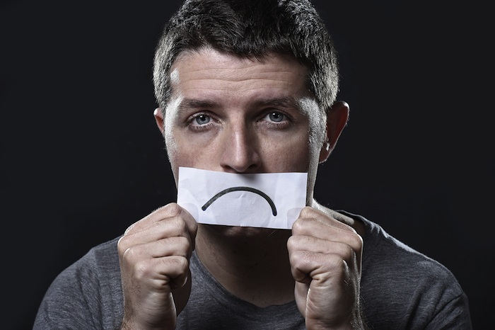 young depressed man lost in sadness and sorrow holding paper with sad mouth draw on his mouth in stress grief depression and lost of hope concept isolated on black background