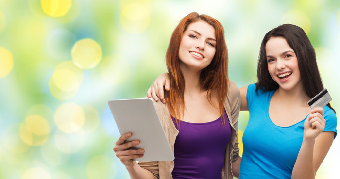 online shopping, e-money, commerce, people and technology concept - two smiling teenage girls or young women with tablet pc computer and credit card over green lights background