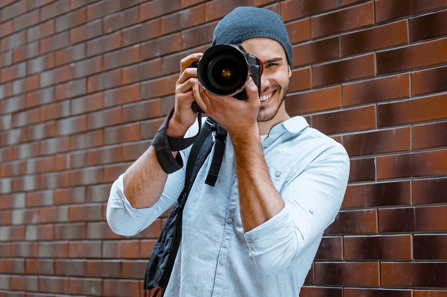 Portrait of stylish handsome young photographer with bristle standing outdoors and leaning on brick wall. Young man wearing shirt and hat. Man with professional camera looking through lens
